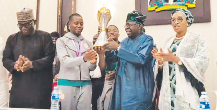 President Bola Tinubu (2nd right), receiving the Trophy from the Captain of the victorious Super Falcons, Rasheedat Ajibade when the President hosted the Nigeria team to the Women’s Africa Cup of Nations (WAFCON) championship in Morocco, yesterday, at Abuja, while his wife, Remi and others applaud the gallant ladies.